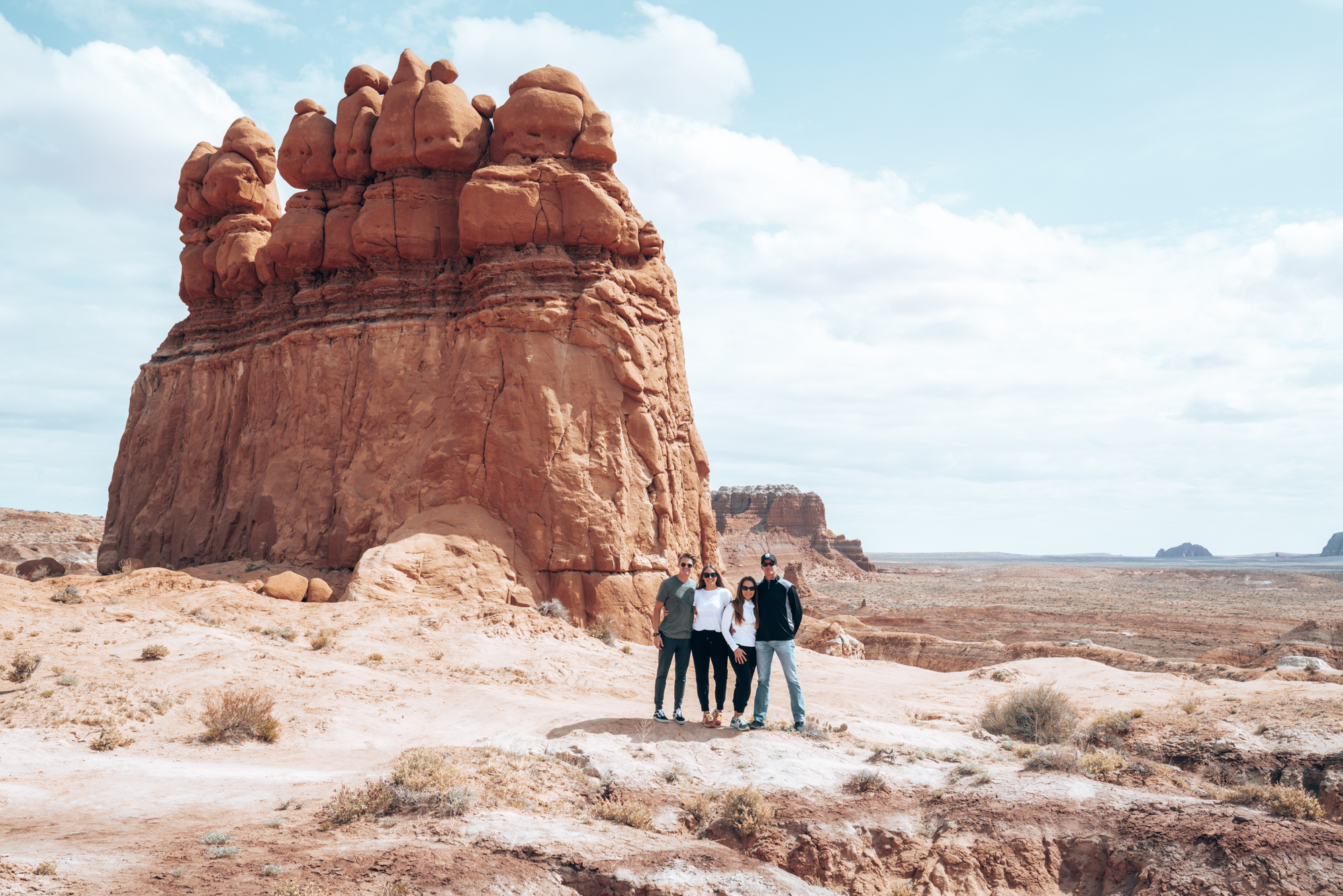 Two couples pose in front of a large red butte with the blue skies behind them. 