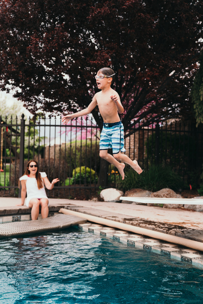 A little boy jumps off a diving board in a backyard swimming pool.
