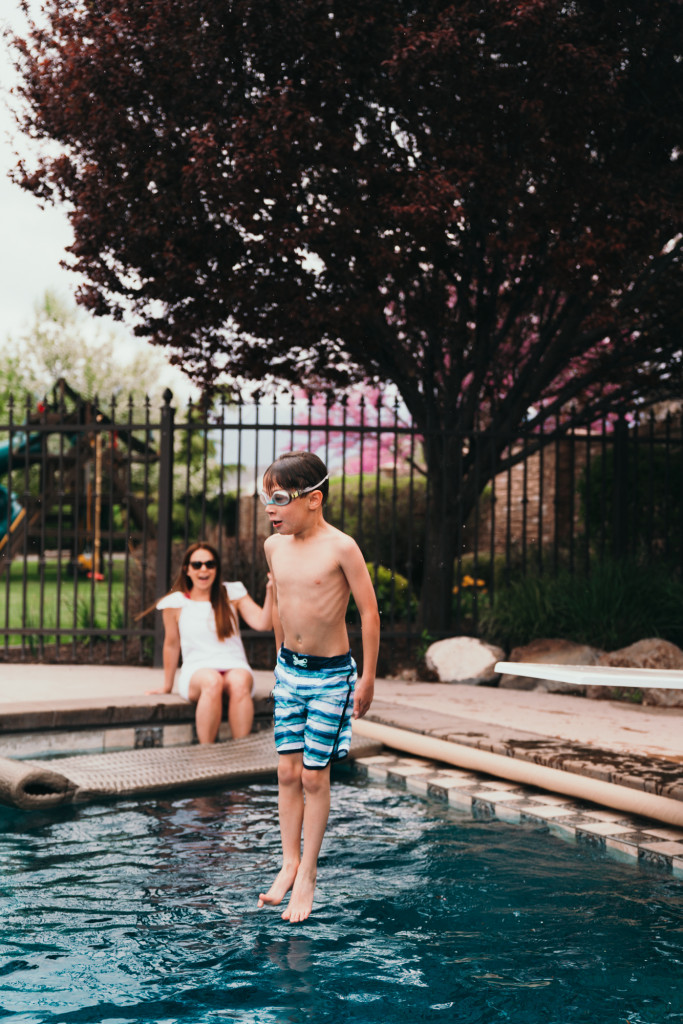 A little boy jumps off a diving board in a backyard swimming pool.