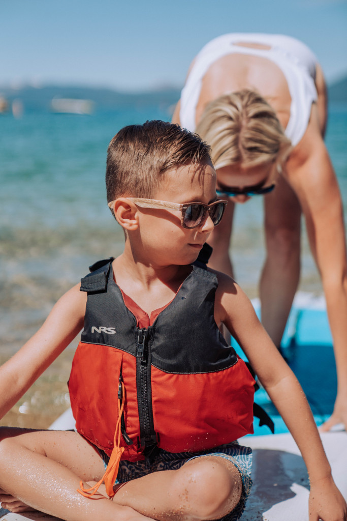 a little boy sits on the front of a paddle board with a life jacket on