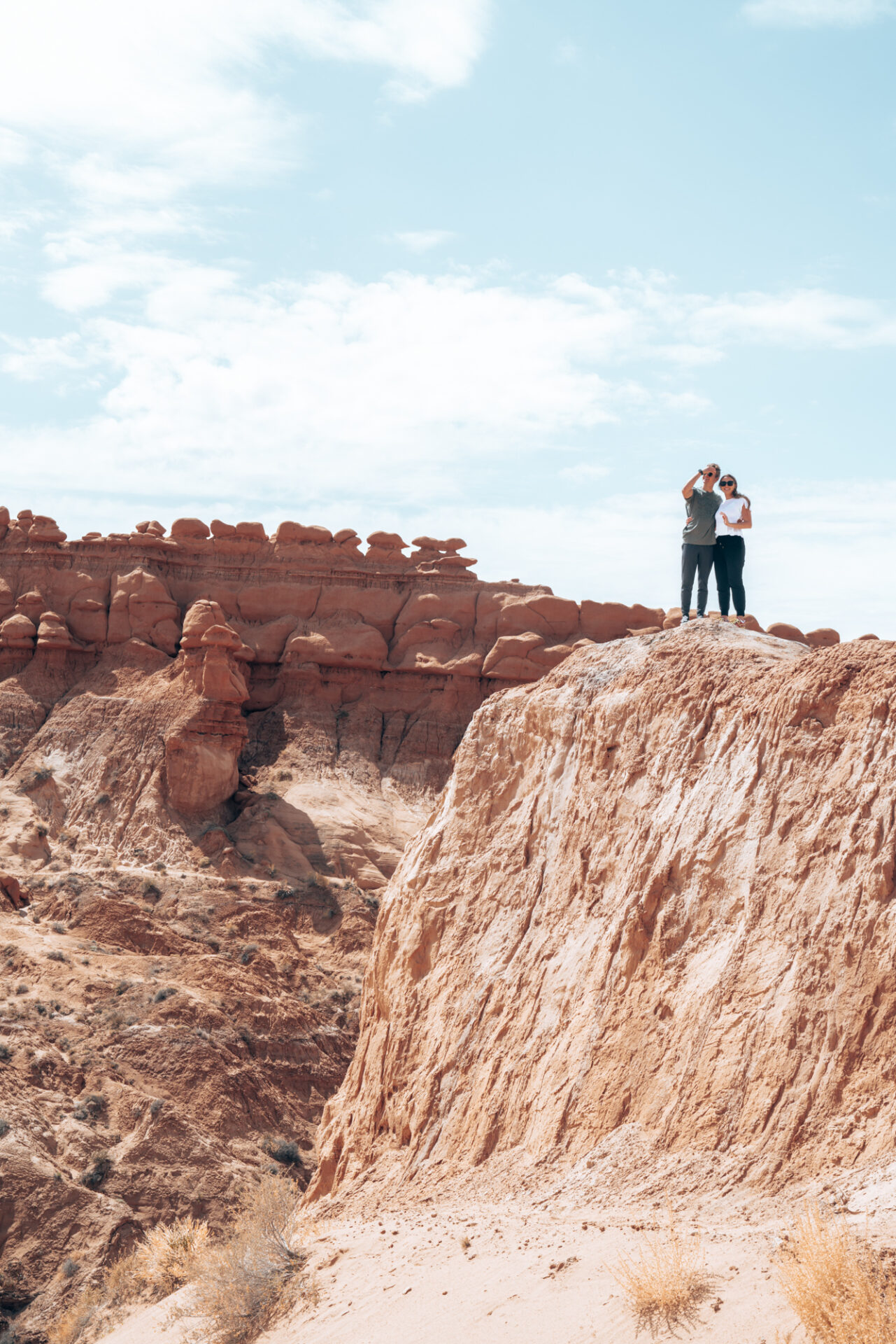 a young couple pose at the edge of cliff with red rock and blue skies behind them.