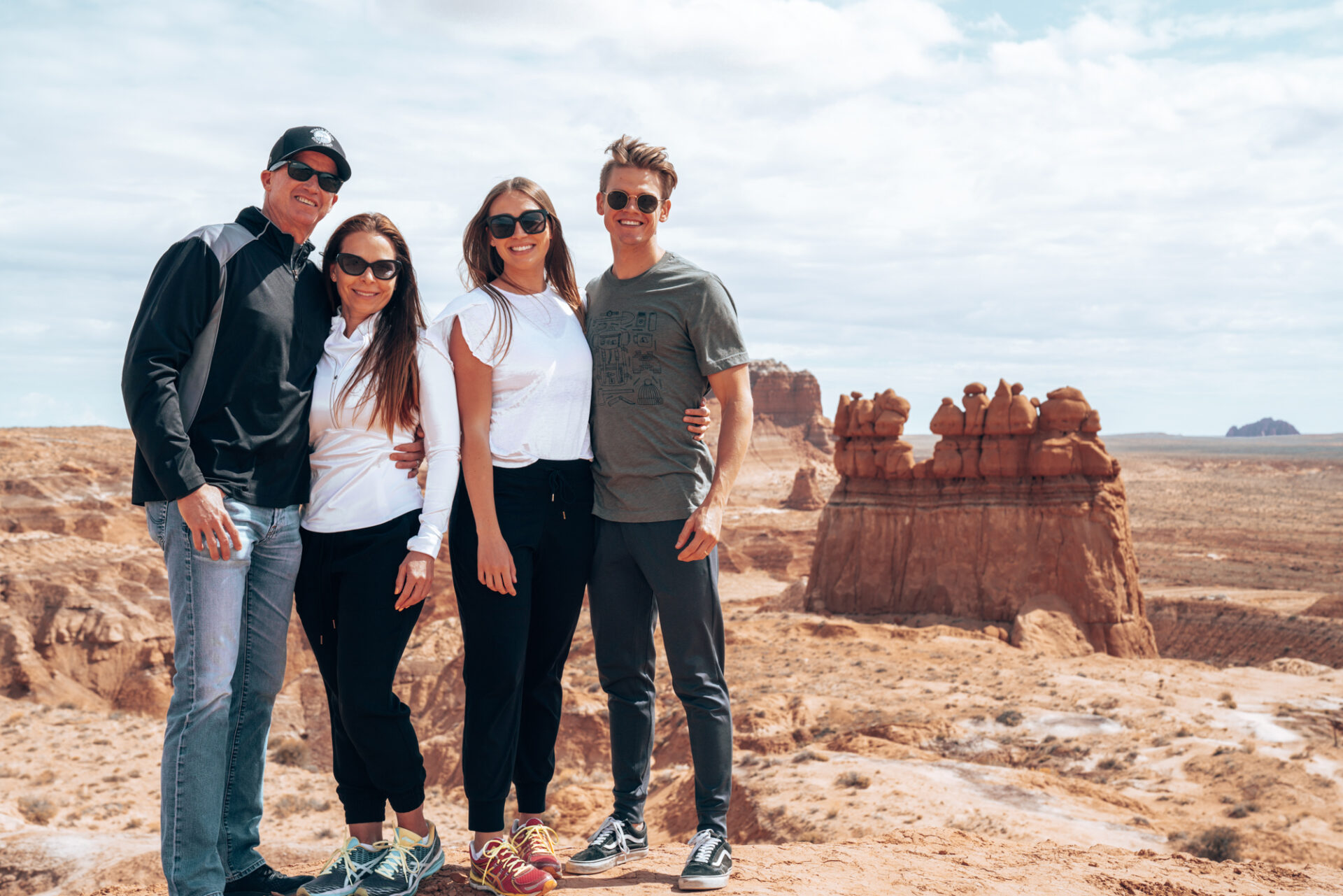 a family of four pose in on a cliff with red rock buttes and blue skies behind them.