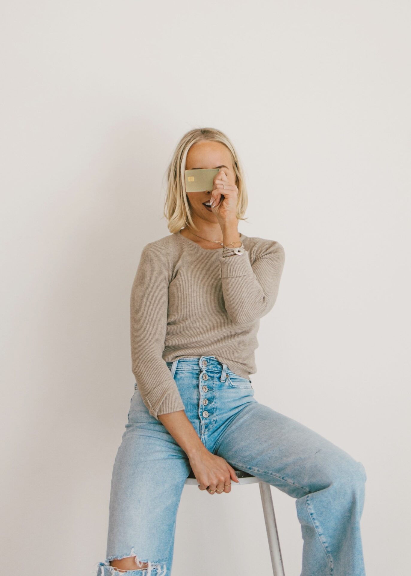 Kam sitting on white stool in beige sweater and distressed jeans smiling