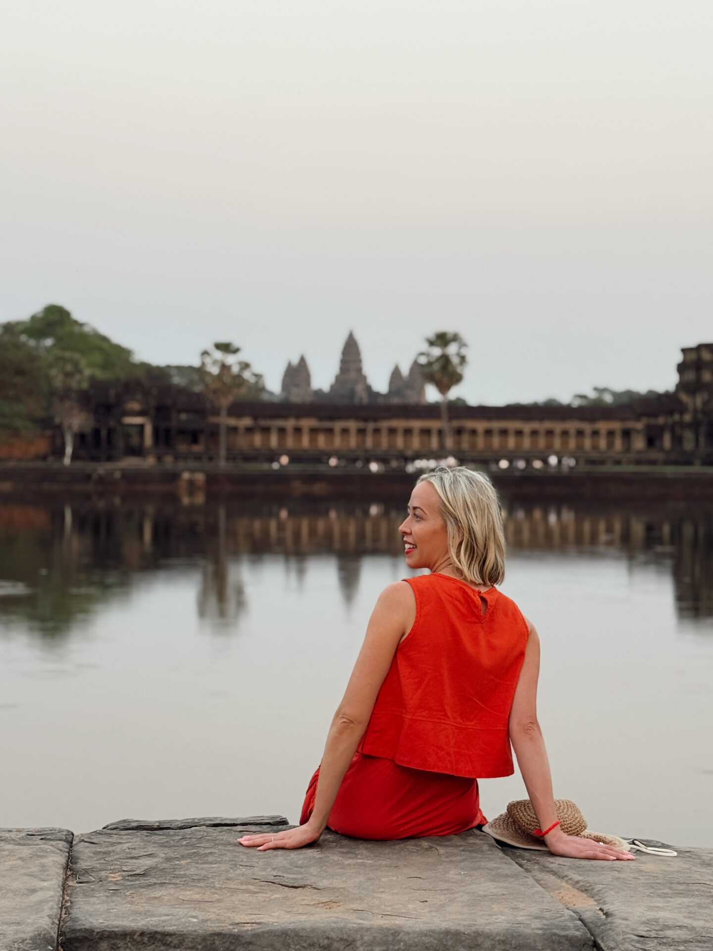 Woman sitting by the moat watching sunrise over Angkor Wat silhouette reflected in the water