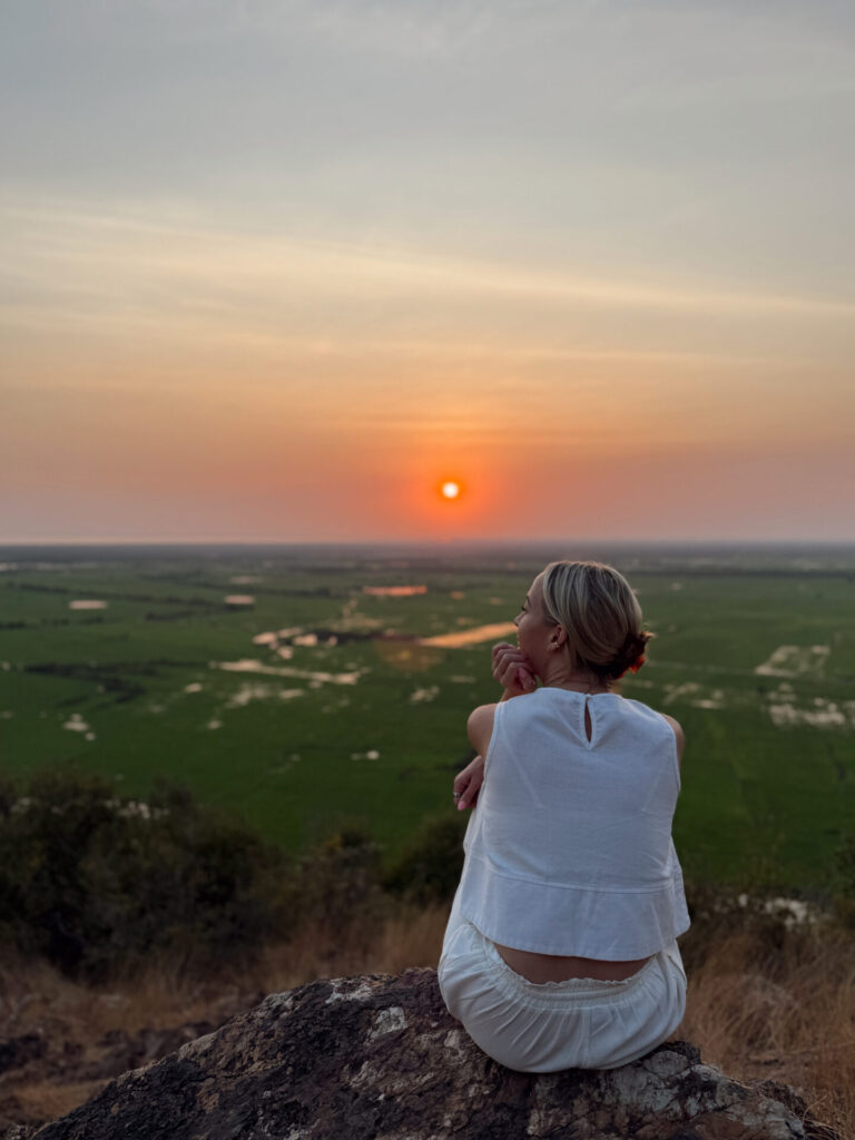 Woman sitting on a rock watching sunset over the green Cambodian countryside from Phnom Krom hill