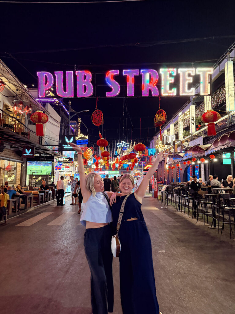 Two friends posing under the illuminated Pub Street sign in Siem Reap at night with red lanterns