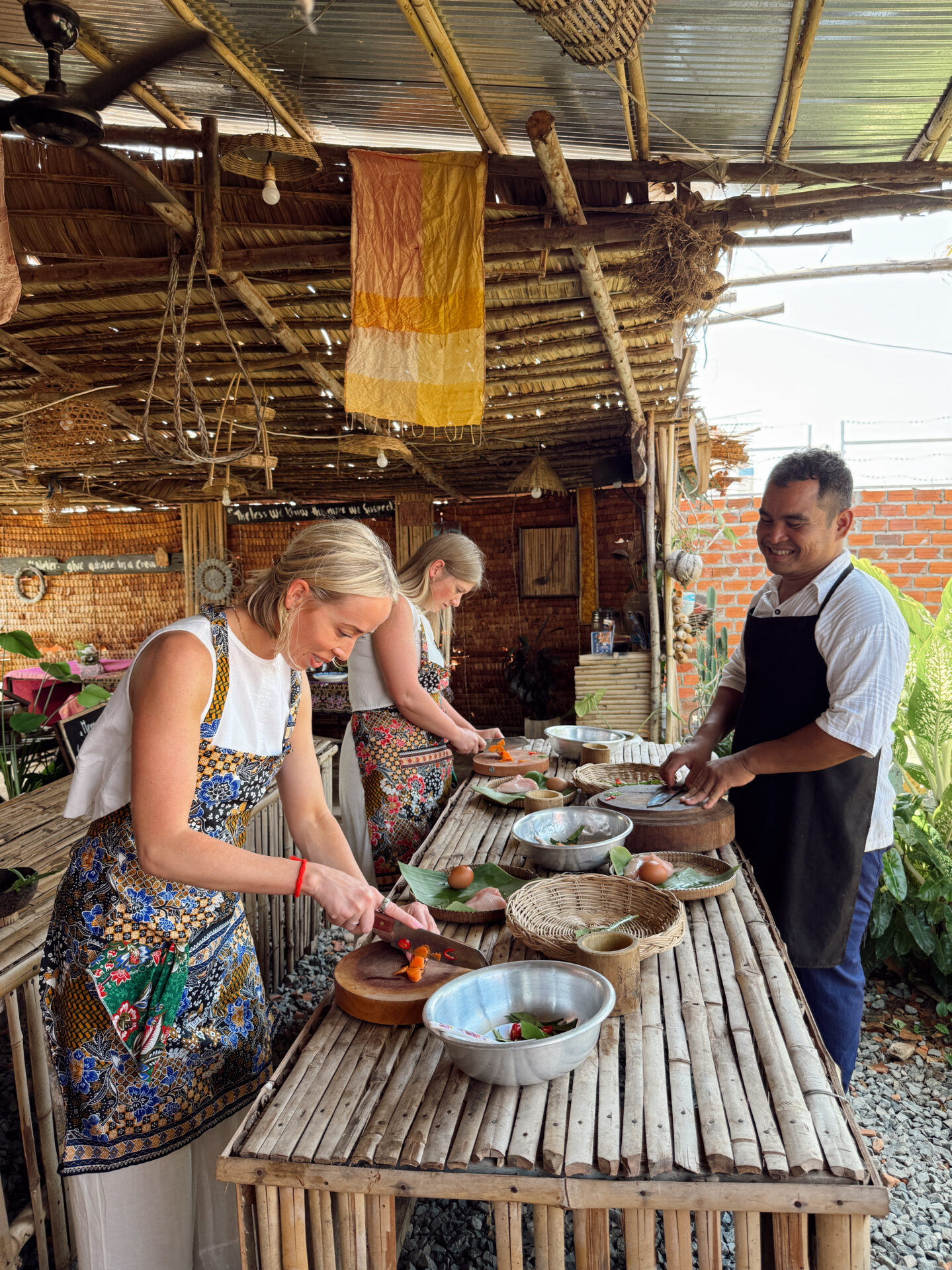Two women and a chef chopping ingredients at a bamboo outdoor kitchen during a Siem Reap cooking class