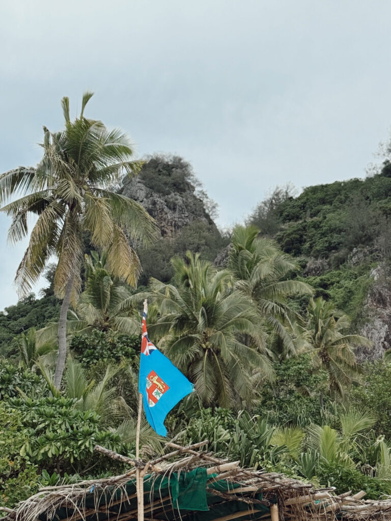 Fiji flag flying from a bamboo raft below a dramatic limestone peak and palm trees