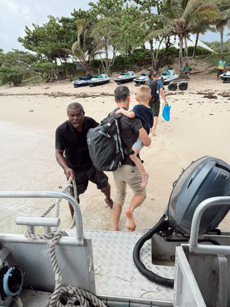 Family boarding a boat transfer from a Fiji beach with jet skis lined up along the sand