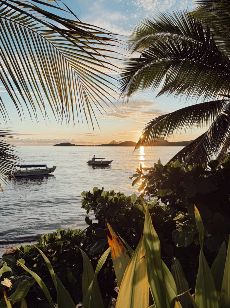 Golden Fiji sunset over calm water with boats anchored offshore, framed by palm fronds