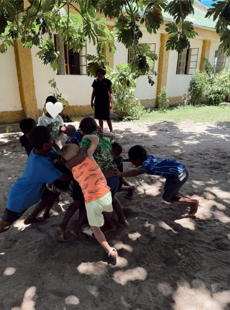 Local Fijian village school children and a visiting boy playing rugby together in a sandy schoolyard