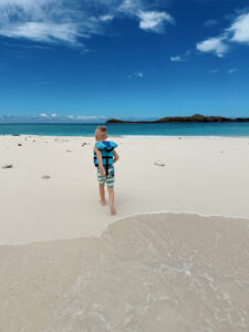 Young boy in a blue life vest walking across a white sandbar toward turquoise water in Fiji