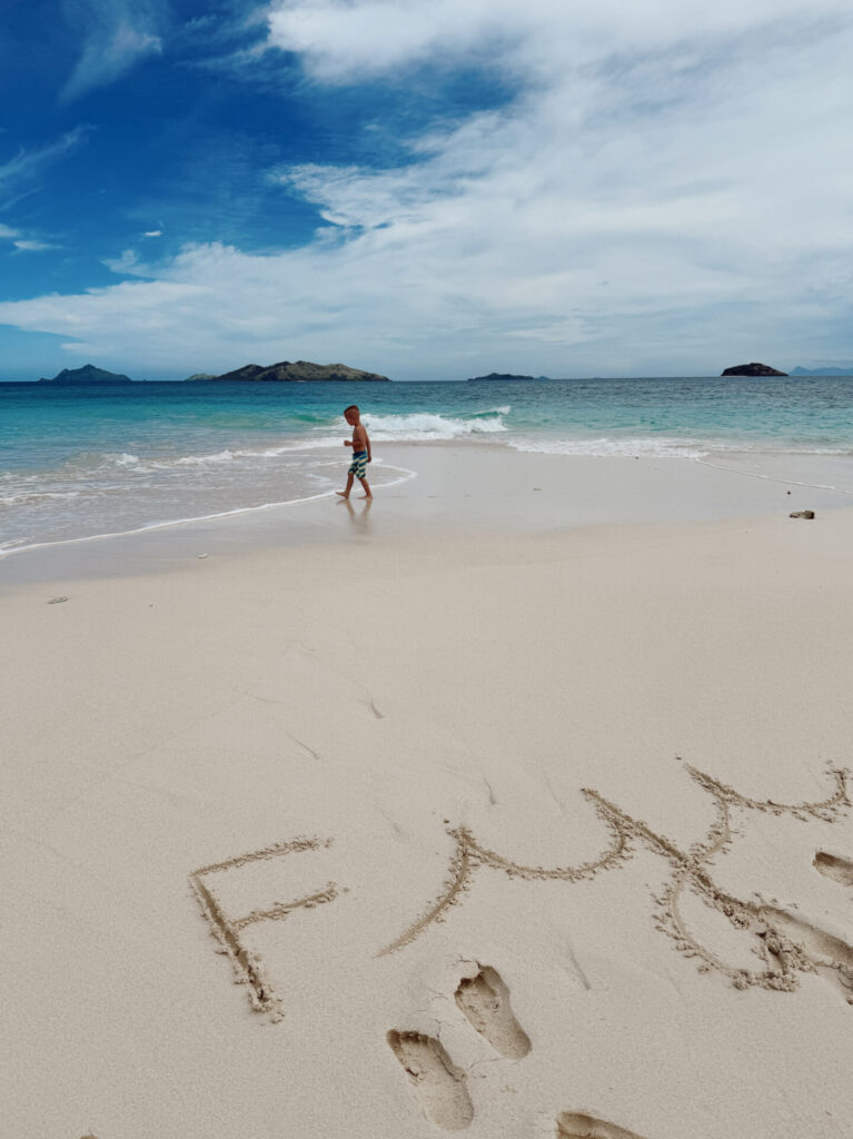 The word Fiji written in white sand on an empty beach as a boy walks near the turquoise shoreline