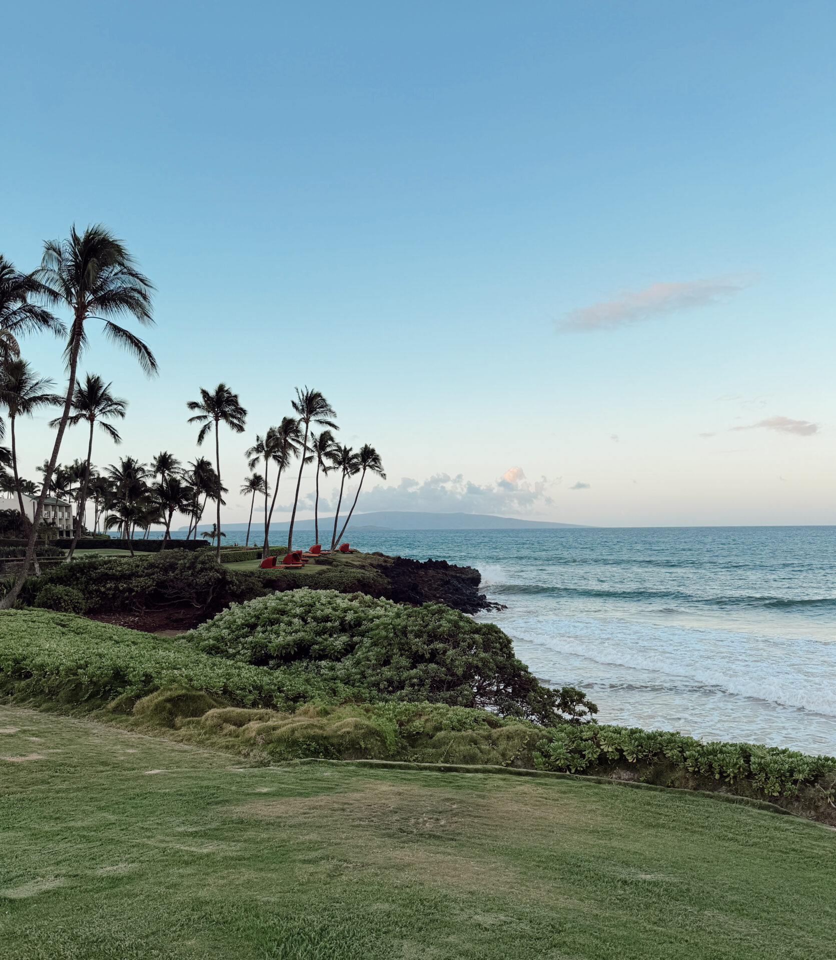 Oceanfront lawn with red lounge chairs and palm trees overlooking Maui's coastline at sunset