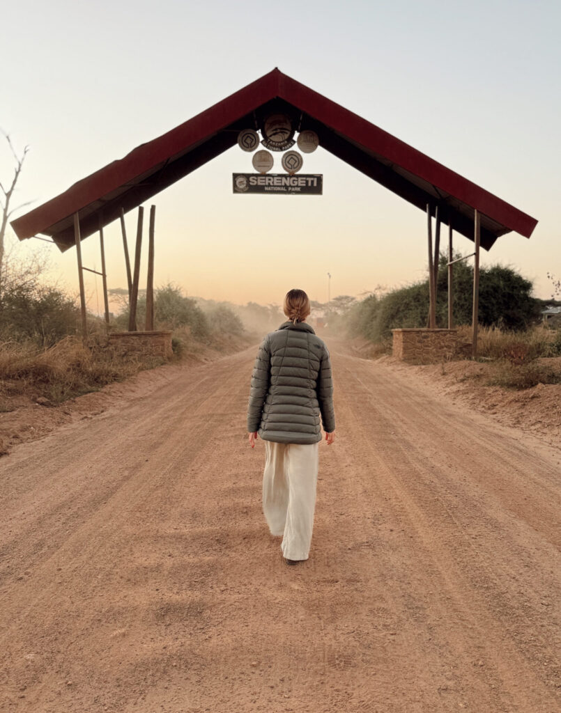 Woman walking toward the Serengeti National Park entrance gate on a dusty road at sunrise
